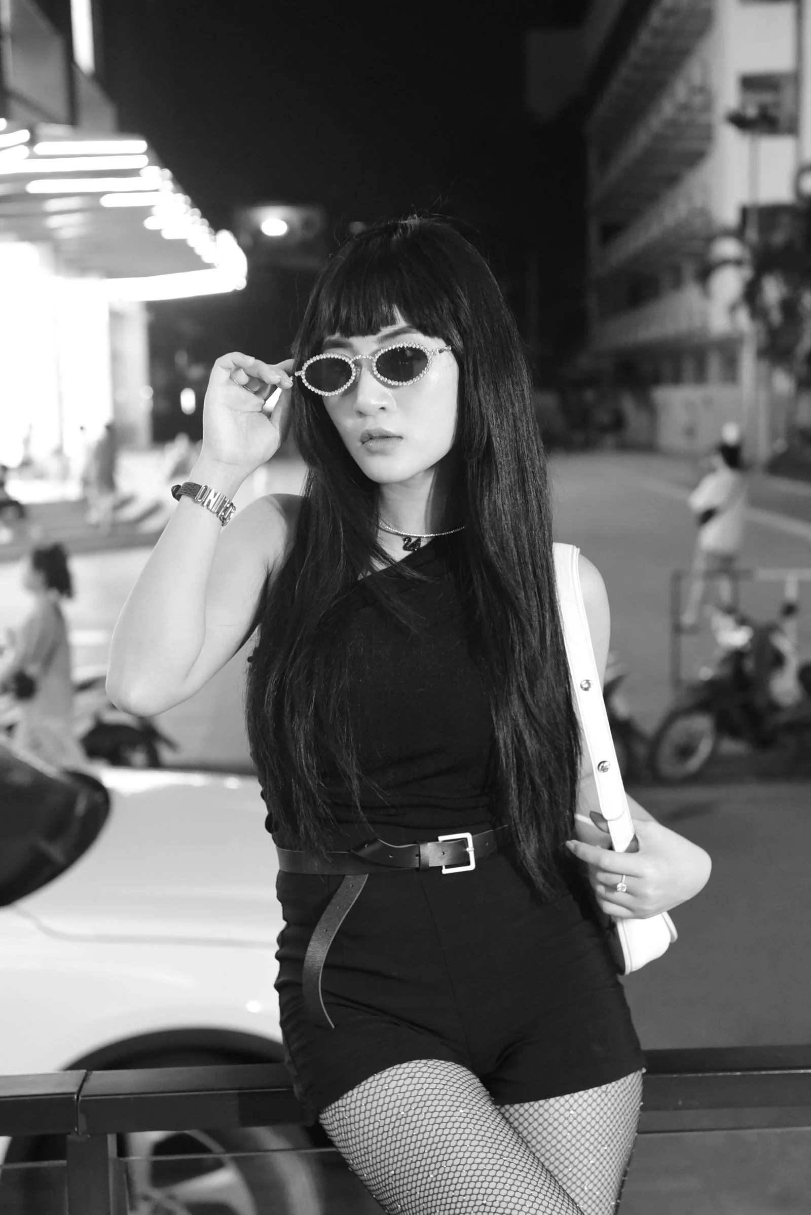 Black and white portrait of a fashionable young woman posing at night in an urban setting with sunglasses and long hair.